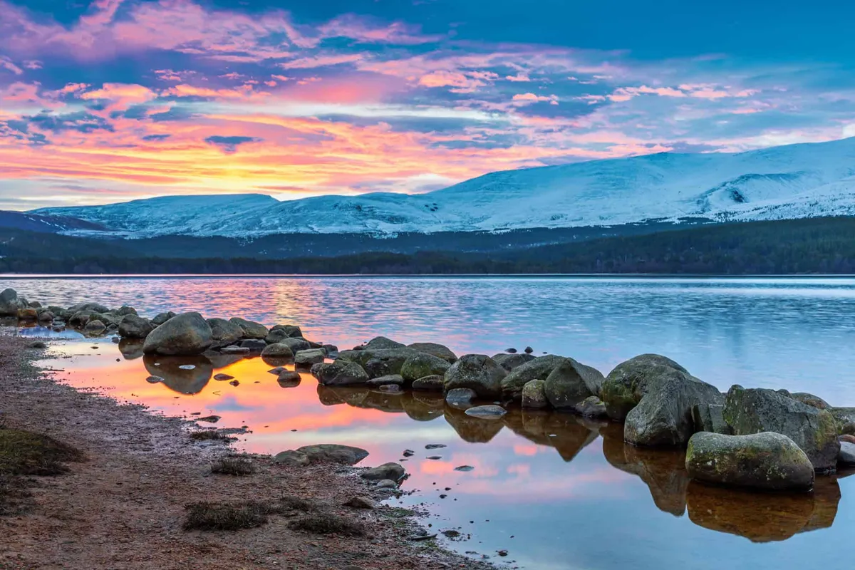 Vibrant pink sunrise reflecting in Loch Morlich with snow-capped Cairngorm mountains in the background.