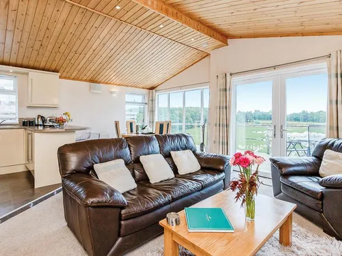 Open-plan living room with leather sofas, pine-clad ceiling, and meadowland views through patio doors