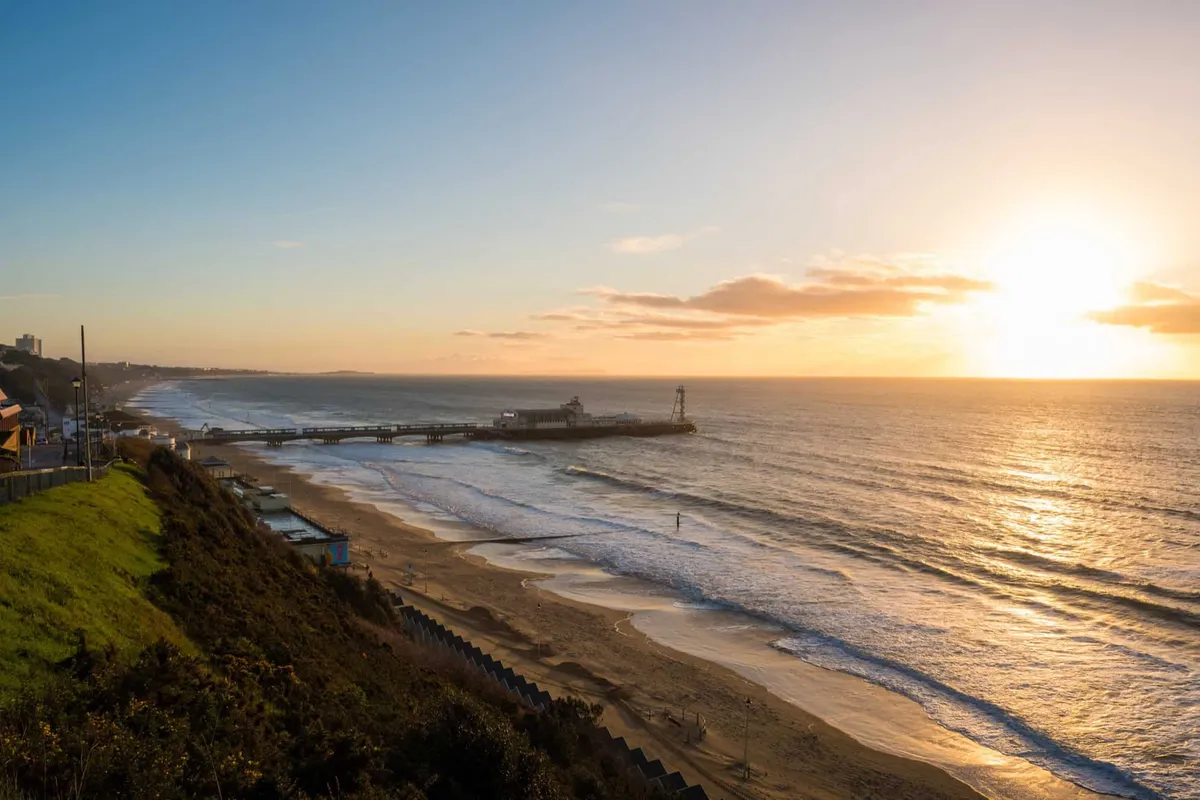 Aerial view of Bournemouth Pier and the golden sandy beach at sunrise, with warm light reflecting off the calm sea.