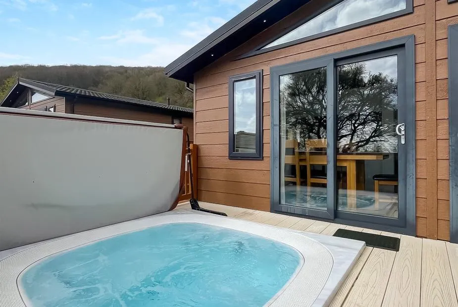Hot tub on decking beside Brunel Premier Elegance Lodge 3 with wooded hillside beyond