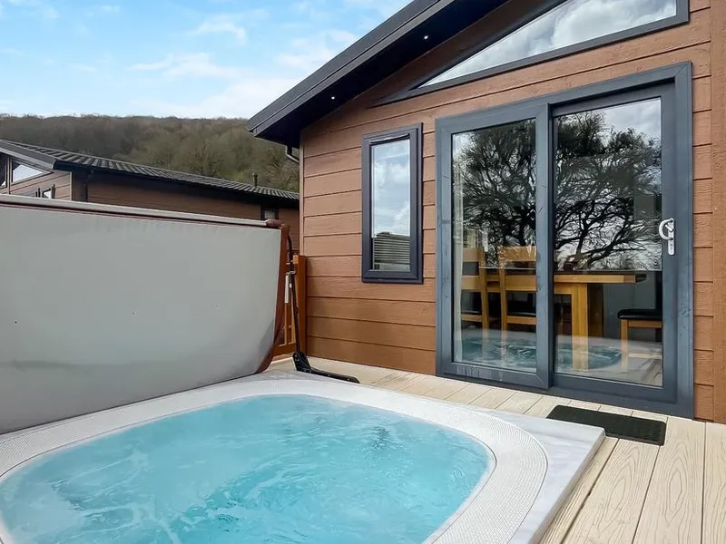 Hot tub on decking beside Brunel Premier Elegance Lodge 3 with wooded hillside beyond