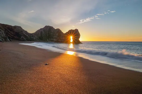 Golden sunrise at Durdle Door in Dorset, featuring a dramatic sunburst shining through the iconic limestone arch onto the sandy beach.