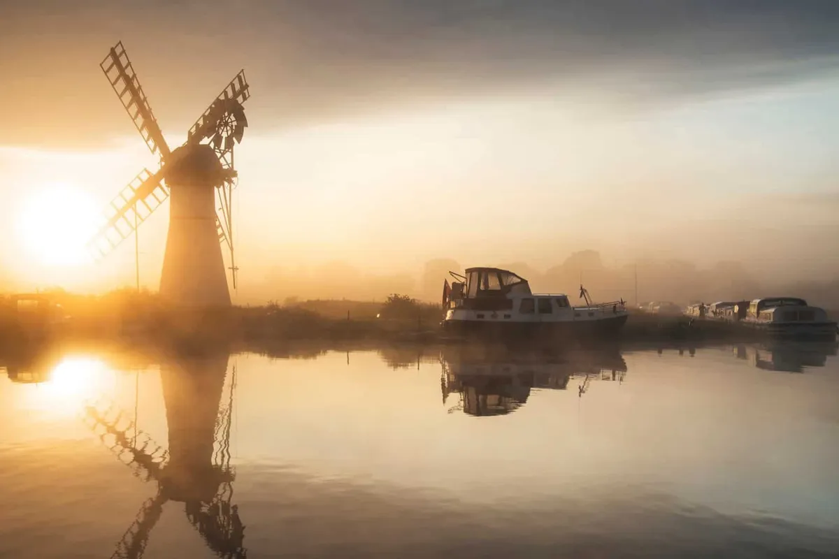 Golden sunrise mist rising over the calm river and Thurne Mill windmill in the Norfolk Broads, East of England.