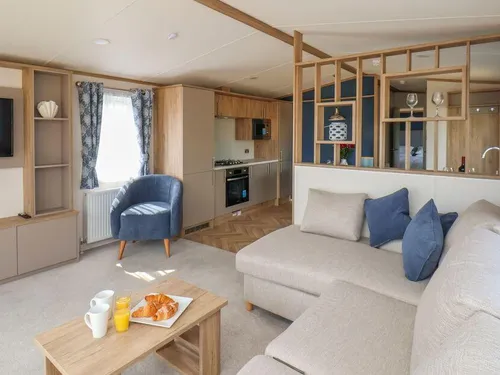 Open-plan living area with corner sofa, blue armchair, wall-mounted TV, and kitchen beyond