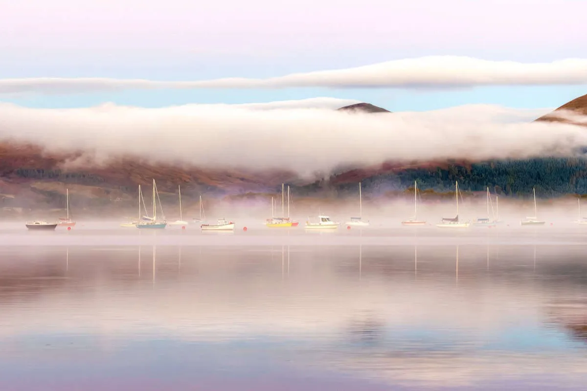 Pastel sunrise with mist rising over the calm waters and moored boats at Milarrochy Bay, Loch Lomond.