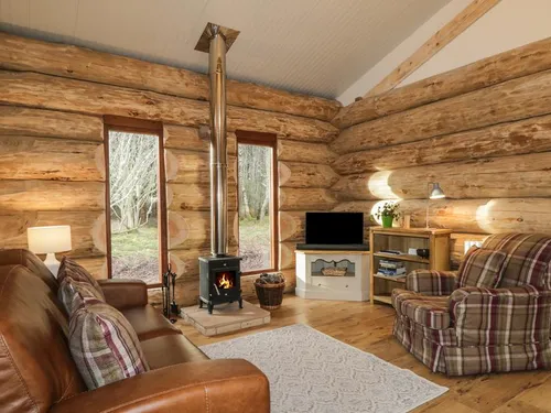 Log cabin living room with wood burner, leather sofa, and tartan armchairs looking out to birch trees