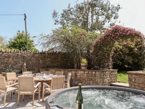 Hot tub on the patio with wooden dining furniture and a flowering pink climbing plant behind stone walls