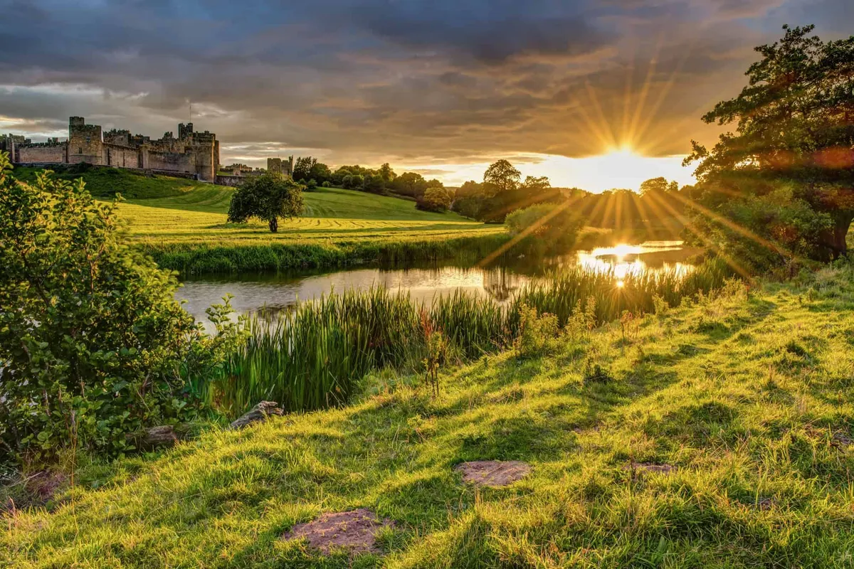Golden sunbeams bursting over the tranquil River Aln and majestic Alnwick Castle in Northumberland.