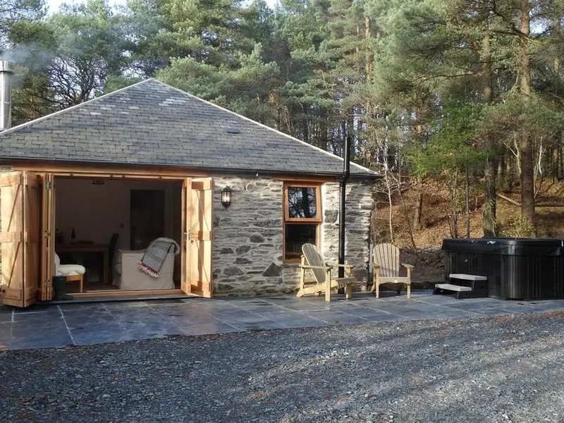 Rock View Cottage with stone exterior, open doors, patio seating and outdoor hot tub in woodland.