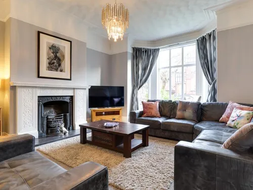 Sitting room with corner sofa, ornate fireplace surround, tall bay windows with curtains, and chandelier
