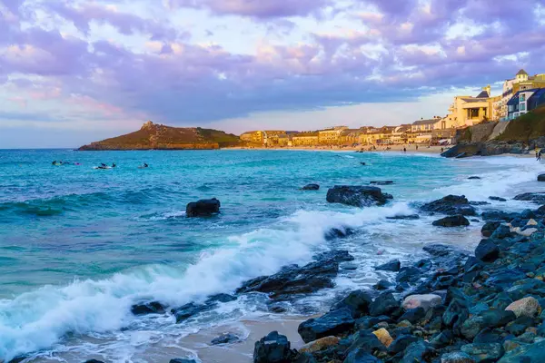 Sunset over Porthmeor Beach in St Ives, featuring golden sands, surfers in the waves, and the iconic headland.