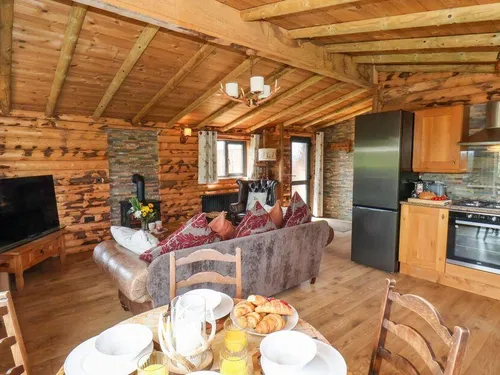 Open-plan living area with log-beamed ceiling, velvet sofa, electric fire, and oak kitchen