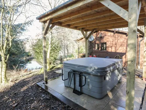 Covered hot tub beside lodge with trees and a small lake in the background.