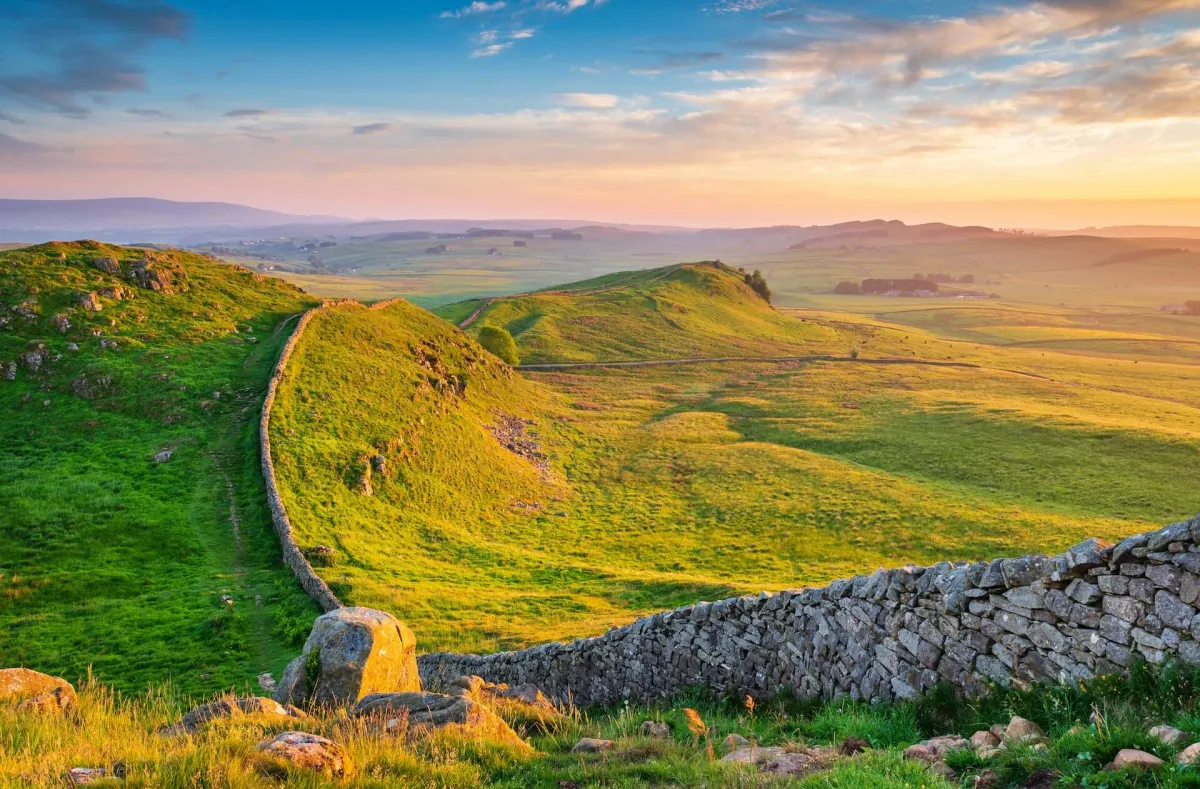 Golden hour sunset over Hadrian's Wall in Northumberland National Park, featuring rolling green hills and historic stone architecture.